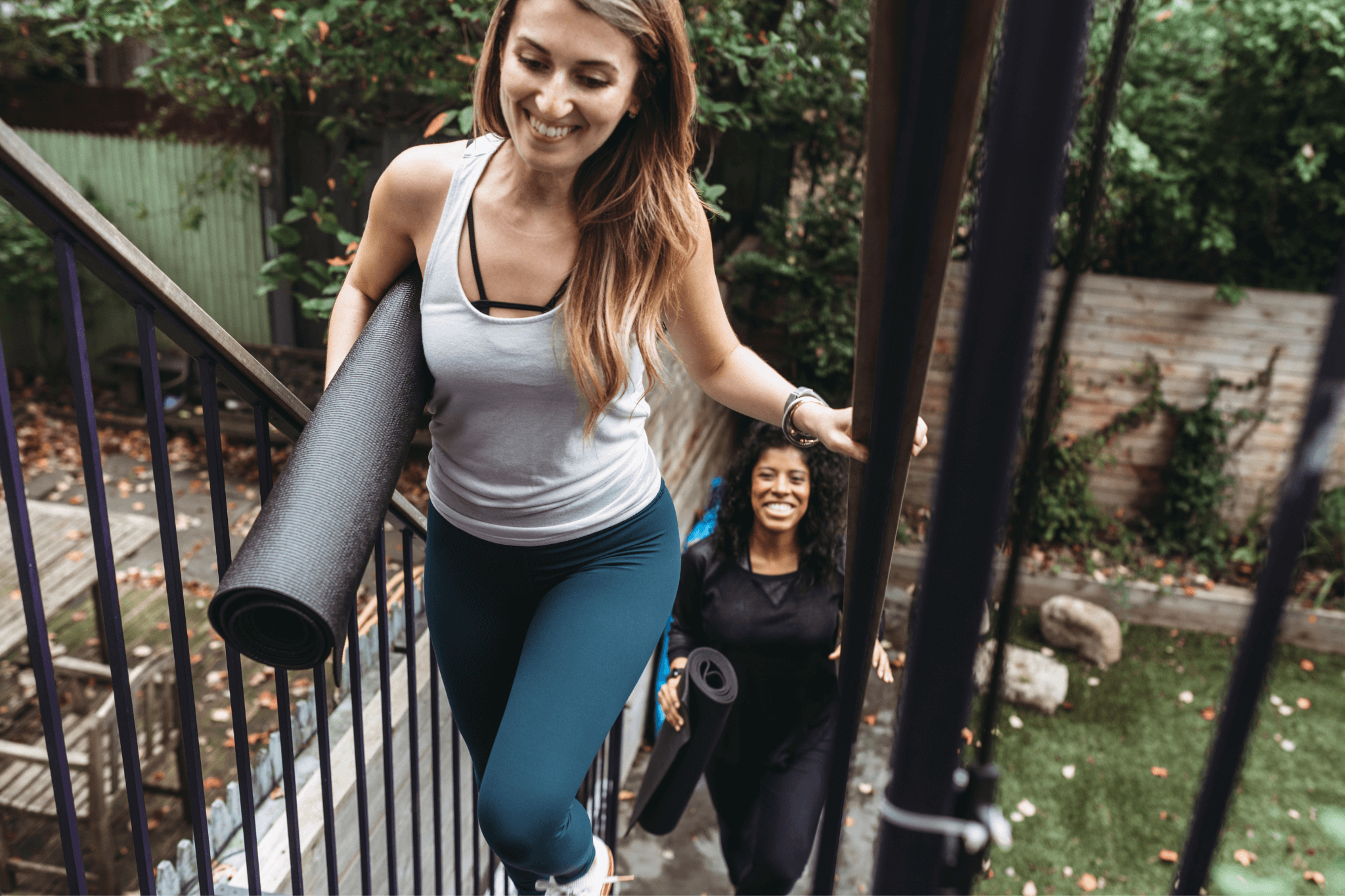 Two women walking up outdoor stairs holding yoga mats, smiling on their way to a yoga session.