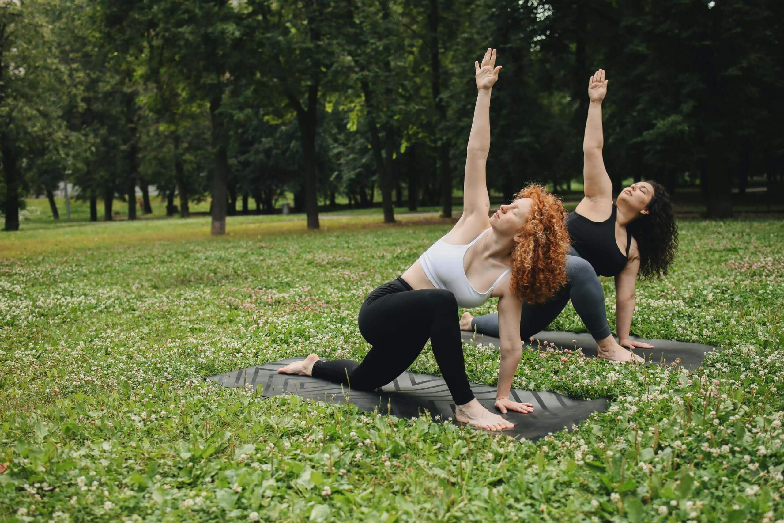 Two women practicing yoga outdoors on mats, holding a deep stretch pose in a green park.